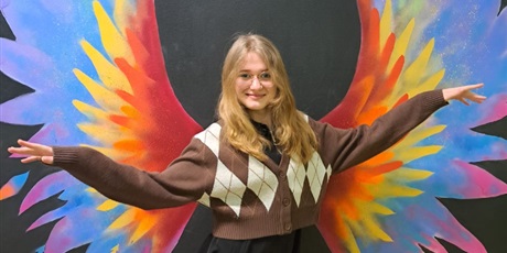 Powiększ grafikę: Dziewczyna w brązowym swetrze pozuje na tle ściany, na której namalowane są kolorowe skrzydła.