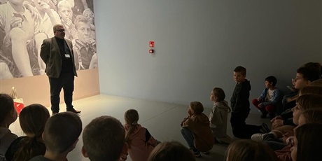 Powiększ grafikę: Przewodnik, a za nim na ścianie zdjęcie ludności z czasów II Wojny Światowej. Uczniowie siedzą i słuchają przewodnika.