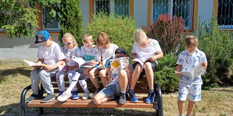 Powiększ grafikę: Uczniowie ubrani na biało siedzą na ławce przed szkołą i czytają książki.