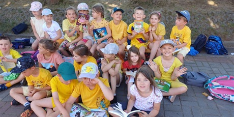Powiększ grafikę: Uczniowie ubrani na żółto siedzą przed szkołą i czytają książki.