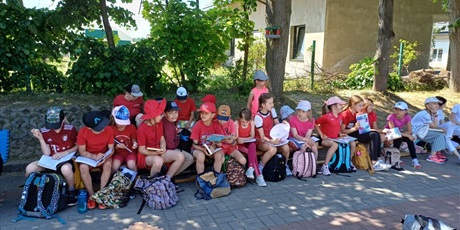 Powiększ grafikę: Uczniowie ubrani na czerwono siedzą przy szkole i czytają książki.