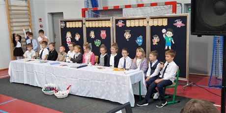 Powiększ grafikę: Uczniowie 2a przebrani za nauczycieli siedzą przy stole symbolizującym stół w pokoju nauczycielskim i grają swoje role.