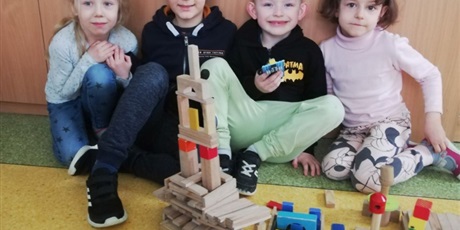 Powiększ grafikę: Czwórka dzieci siedzi na podłodze przy swojej budowli z drewnianych klocków.