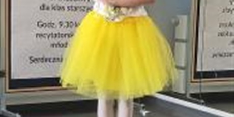 Powiększ grafikę: Dziewczynka w żółtej sukience i białej bluzce stoi przy tablicy dekoracyjnej Miejskiego konkursu recytatorskiego: Brzechwa - Przyjaciel dzieci.