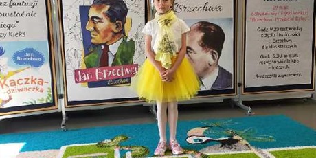 Powiększ grafikę: Dziewczynka w żółtej sukience stoi przy tablicy dekoracyjnej Miejskiego konkursu recytatorskiego: Brzechwa - Przyjaciel dzieci.