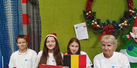 Powiększ grafikę: Pani Barbara Ignaciuk siedzi przy stoisku Unii Europejskiej z dwiema uczennicami i jednym uczniem. Uczennice trzymają flagi państw Europejskich.