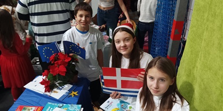 Powiększ grafikę: Dwie uczennice i jeden uczeń siedzą przy stoisku Unii Europejskiej i trzymają flagi państw europejskich.