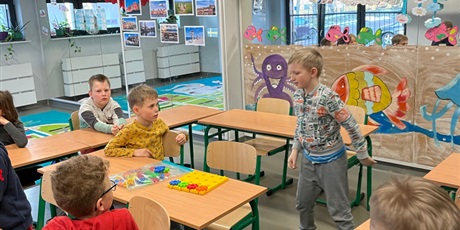 Powiększ grafikę: Dzieci siedzą w klasie przy stolikach parami i w parach kodują z wykorzystaniem klocków KORBO. Jeden chłopiec stoi obok ławki szkolnej.