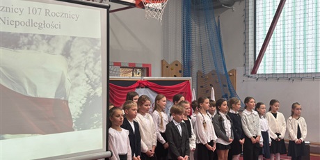 Powiększ grafikę: Klasa 3a śpiewa pieśń patriotyczną. Po jej lewej stronie ekran z wyświetloną flagą Polski. W tle biało-czerwona dekoracja z nazwą Święto Niepodległości 11 listopada.