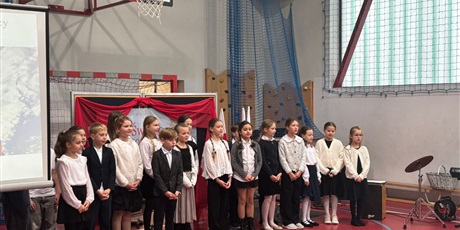 Powiększ grafikę: Klasa 3a śpiewa pieśń patriotyczną. Po jej lewej stronie ekran z wyświetloną flagą Polski. W tle biało-czerwona dekoracja z nazwą Święto Niepodległości 11 listopada.