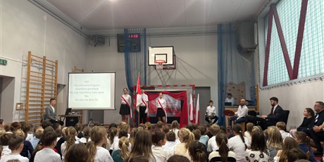 Powiększ grafikę: Widok na scenę. W centrum poczet sztandarowy. Po lewej stronie ekran z tekstem pieśni patriotycznej. Naprzeciwko sceny publiczność siedzi.