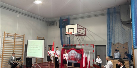 Powiększ grafikę: Widok na scenę. W centrum poczet sztandarowy. Po lewej stronie ekran z tekstem pieśni patriotycznej. 