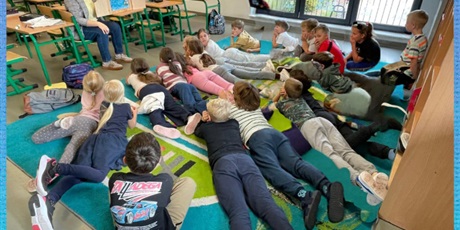 Powiększ grafikę: Bibliotekarka siedzi przy stoliku i czyta opowieść w Teatrzyku Kamishibai. Dzieci leżą przed Pania na dywanie i słuchają. 