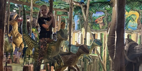 Powiększ grafikę: Dziewczynka na karuzeli siedzi na dinozaurze.