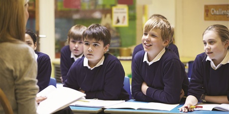 Powiększ grafikę: Nauczyciel siedzi naprzeciwko ławek wypełnionych uczniami i im czyta.