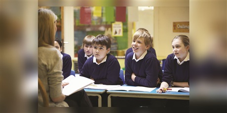 Powiększ grafikę: Nauczyciel siedzi naprzeciwko ławek wypełnionych uczniami i im czyta.