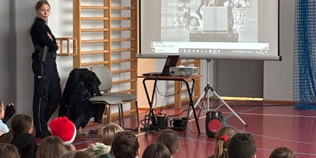 Powiększ grafikę: Pan policjantka prezentuje uczniom film o bezpieczeństwie na ekranie z rzutnika. Jest z dziećmi na sali gimnastycznej.