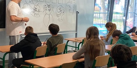 Powiększ grafikę: Ucziowie siedzą w ławkach i pod kierunkiem prowadzącego warsztaty (który sysuje postacie na tablicy) rysują własne postacie komiksowe.