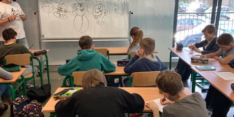 Powiększ grafikę: Ucziowie siedzą w ławkach i pod kierunkiem prowadzącego warsztaty (który sysuje postacie na tablicy) rysują własne postacie komiksowe.